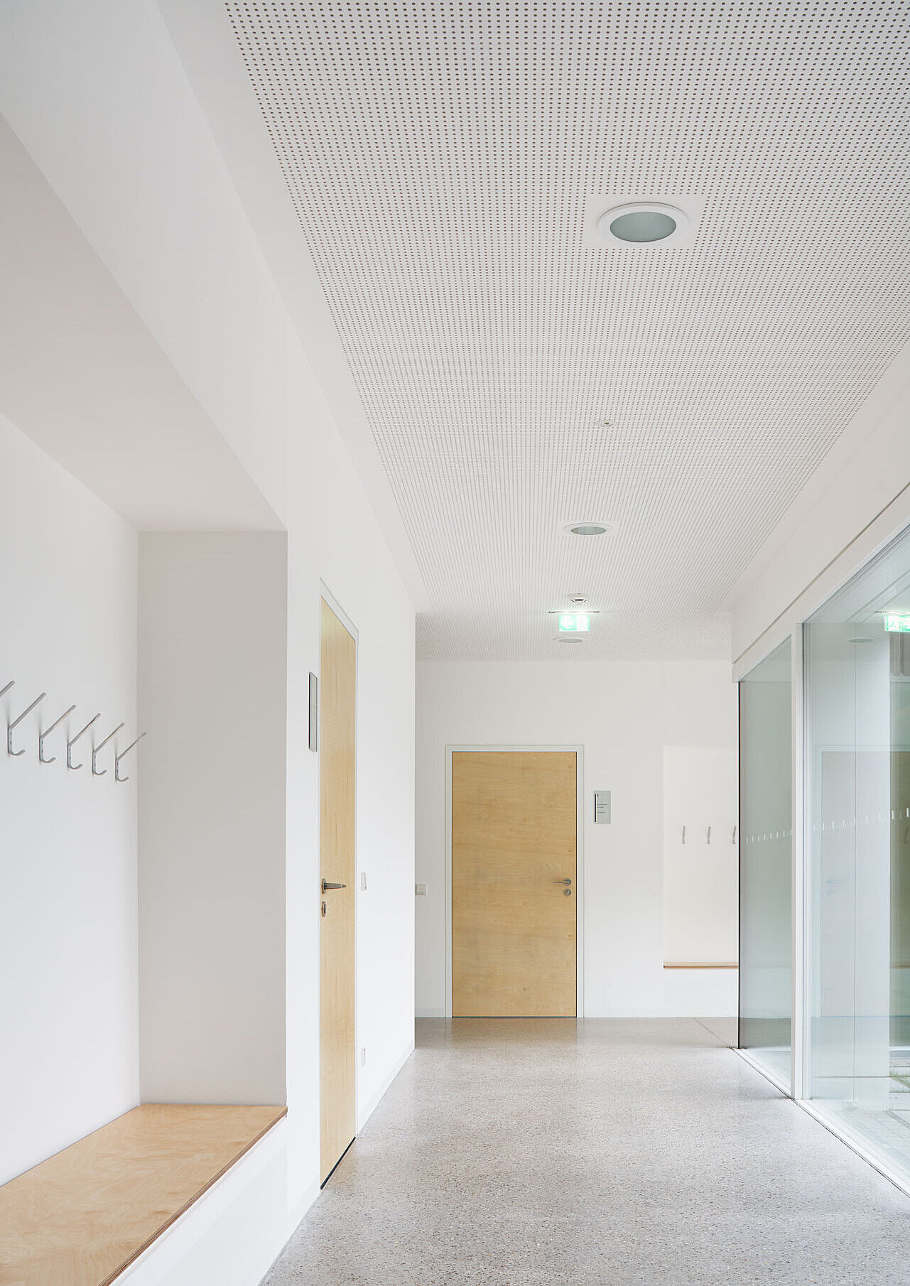 Zu sehen ist der lichtdurchflutete Flur mit weißen Wänden und Holztüren zu den Klassenzimmern, ausgestattet mit Garderobe und Sitzgelegenheiten.