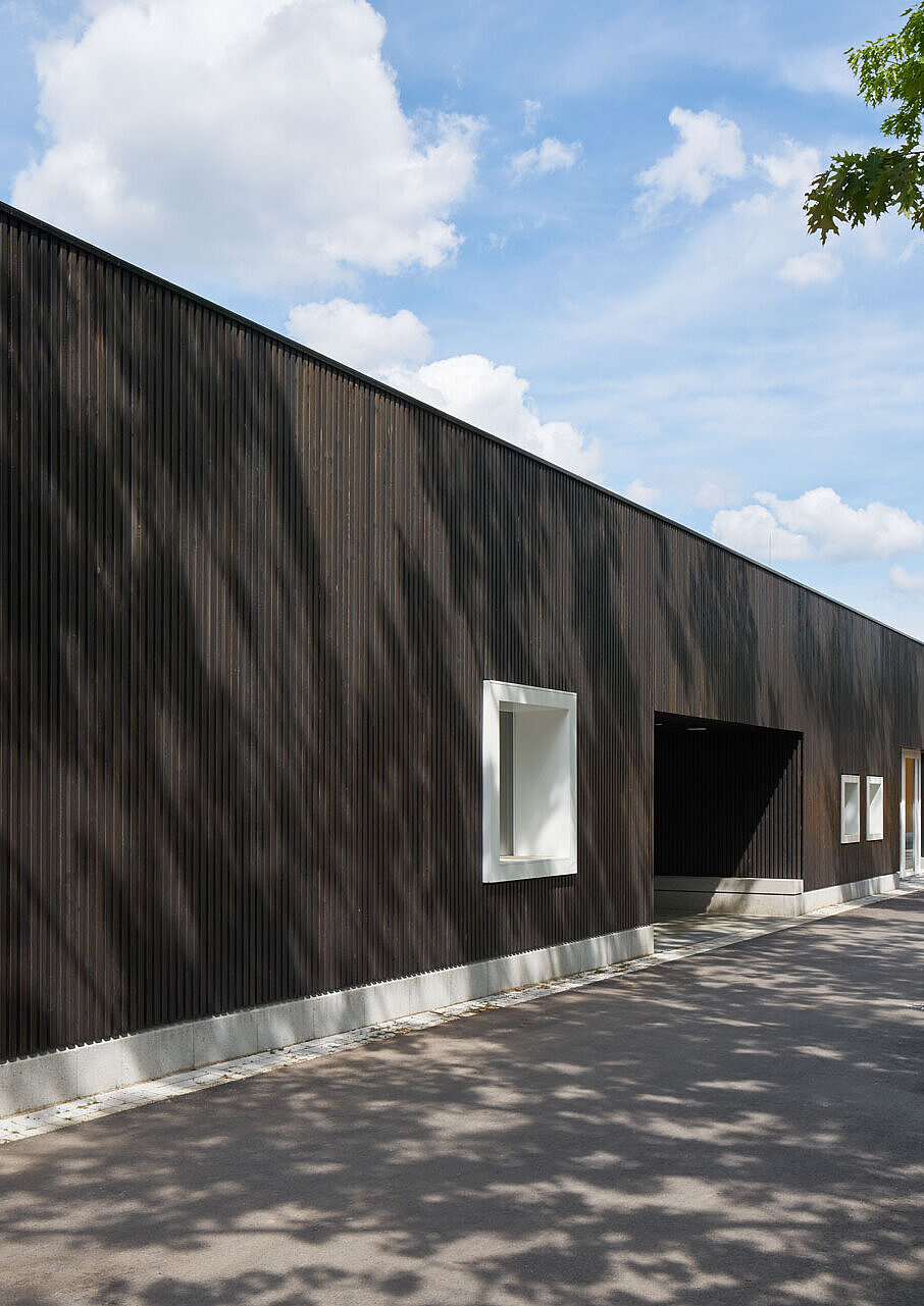 Zu sehen ist die Südwestansicht der grau lasierten Holzlattenfassade mit einem hellgrauen Sockelstein und mehreren Fenstern mit kräftigen weißen Holzrahmen.