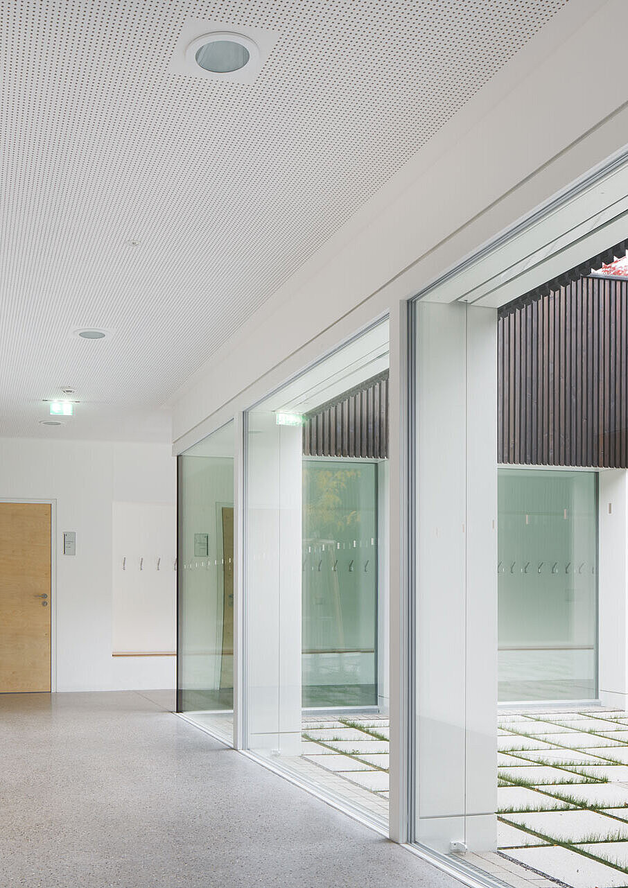 Blick entlang des lichtdurchfluteten Flurs auf eine geschlossene Holztür eines Klassenzimmers, daneben bieten die großen Fenster einen Blick auf den Lichthof.