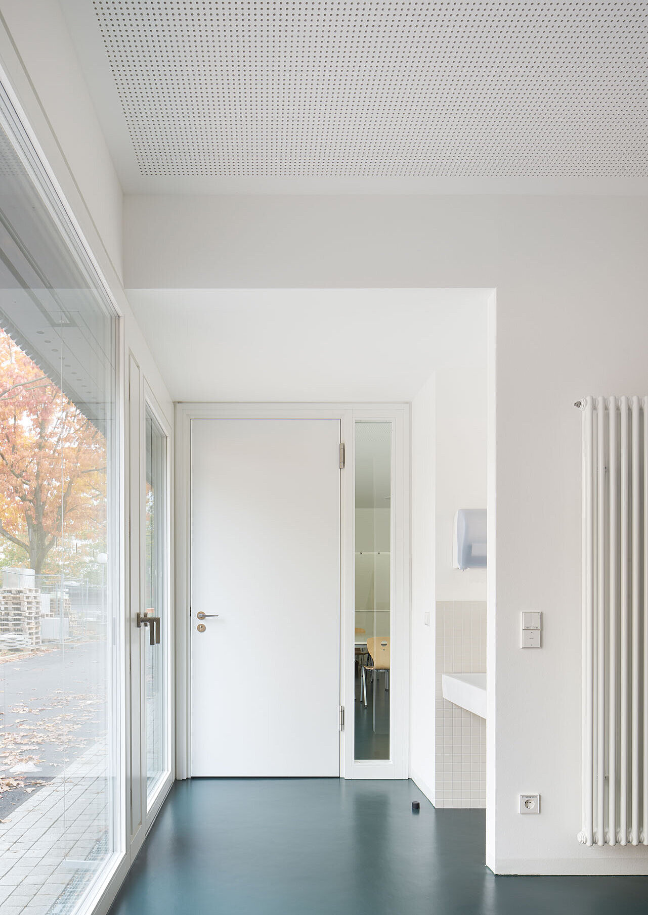 Blick auf die weiße Tür zum Aufenthaltsraum, links daneben befinden sich bodentiefe Fenster mit weißen Rahmen.