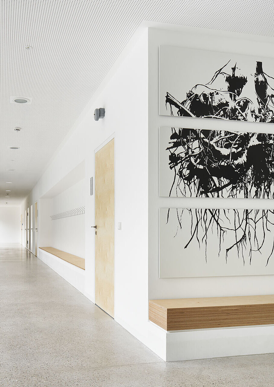 Blick entlang des lichtdurchfluteten Flurs mit weißen Wänden, Kunst am Bau sowie Holzakzenten an Türen und Sitzgelegenheiten.