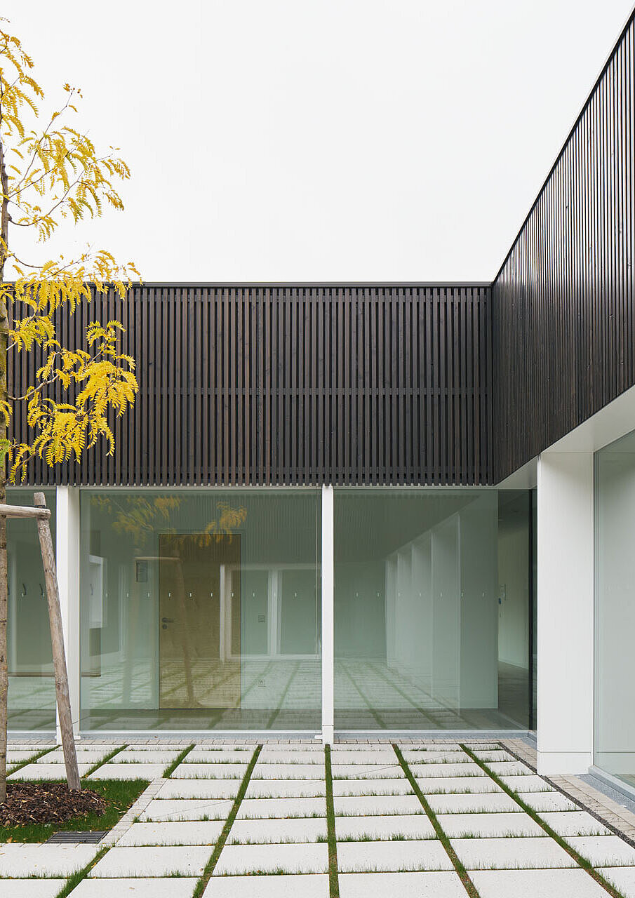 Blick vom Innenhof auf das Rasenpflaster und einen frisch gepflanzten Baum, dahinter ein Teil der dunkelgrauen Holzlattenfassade mit großen Fenstern und weißen Rahmen.