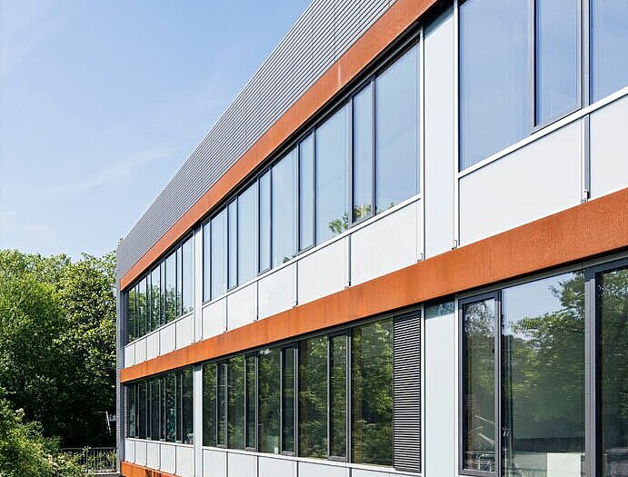  Das Bild zeigt einen Teil der Südperspektive des Gebäudes, bei dem das Zusammenspiel der anthrazitfarbenen und bronzefarbenen Fassade gut zur Geltung kommt.