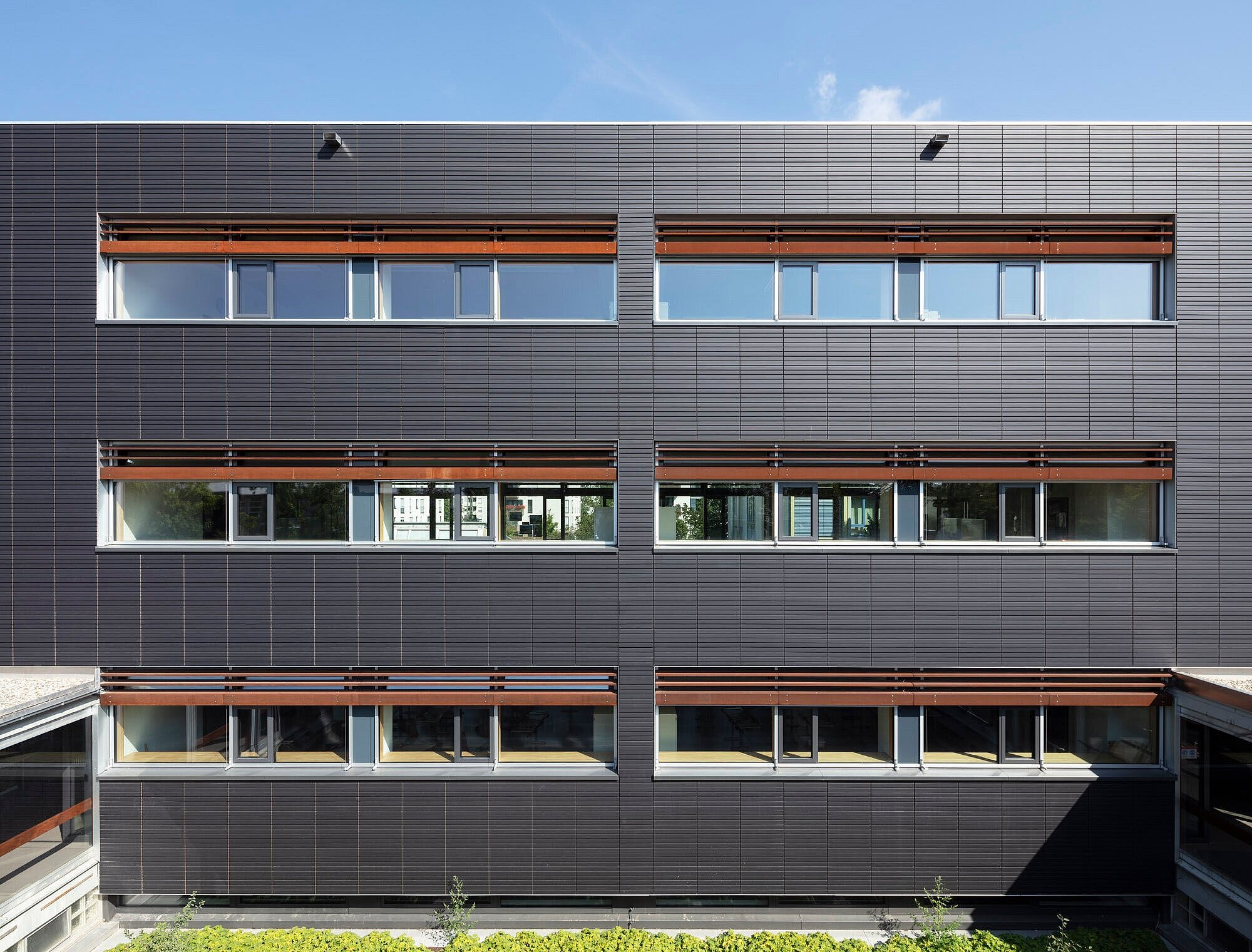 Zu sehen ist ein Teil der anthrazitfarbenen Nordfassade mit Fensterbändern und Blenden aus Cortenstahl.