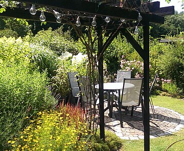 Das Bild zeigt eine Sitzgelegenheit zwischen Stauden und Rasen mit einer Pergola in der Kleingartenanlage SChloss.