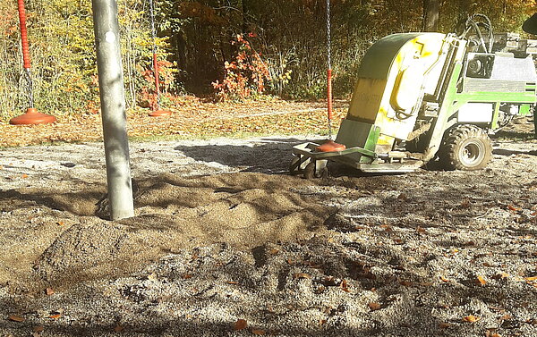 Das Bild zeigt die Reinigung und Lockerung des Fallschutzkieses unter einer einachsigen Überkopfwippe. 