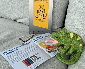 Stoffchamäleon stitzt auf einer Couch und liest Infomaterial eine zusammengeklappte Bille liegt dabei