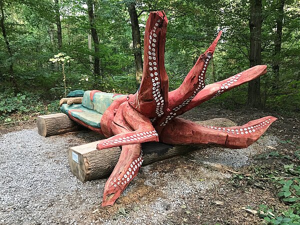 Das Bild zeigt eine Holzbank im Wald in Form einer Krake.