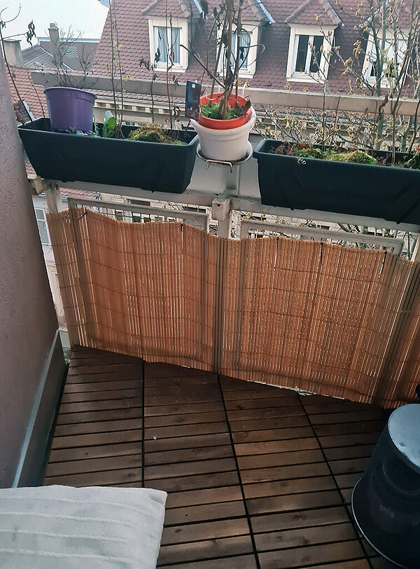 Foto einer Ecke des Balkons mit einem Sichtschutz aus Schilf und einigen Balkonkästen mit verkümmerten Pflanzen.