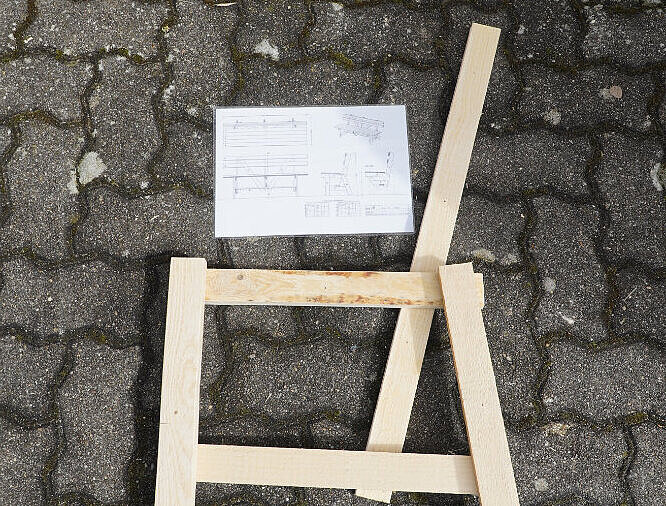 Latten, die angeordnet wie ein Stuhl auf gepflasterten Boden liegen. Darüber eine technische Zeichnung.