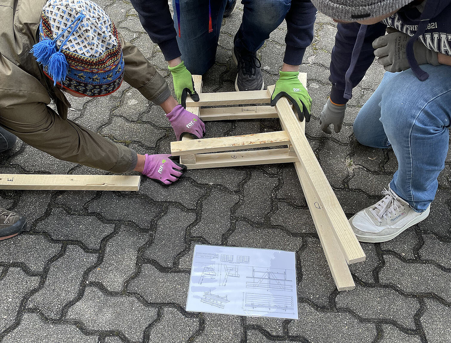 Drei Personen, die arrangierte Holzlatten auf dem Boden halten und auf Plan schauen.