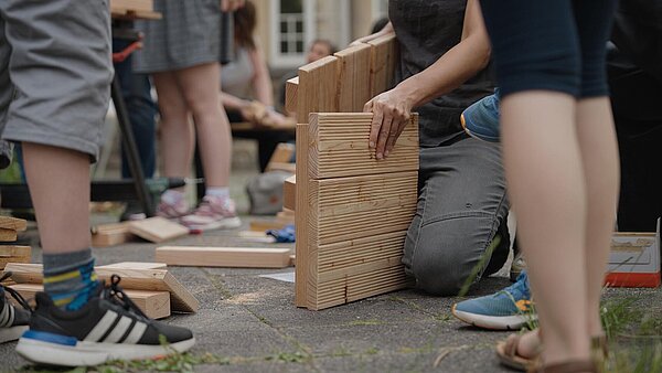 Am Boden knieende Person mit Holzplatten in der Hand. Beine von anderen Leuten drumherum.