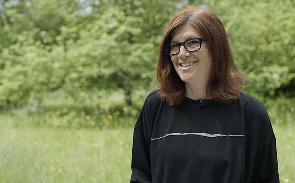 Eine Frau steht lächelnd auf einer Wiese, im Hintergrund Bäume