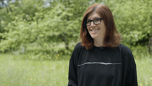 Eine Frau steht lächelnd auf einer Wiese, im Hintergrund Bäume