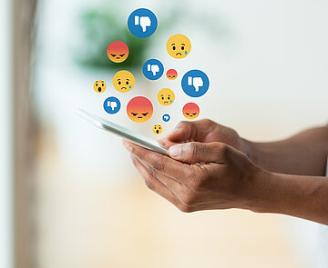 Person hält Handy in den Händen über dem traurige und wütende Emojis zu sehen sind. 