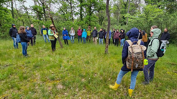 Studierende der Pädagogischen Hochschule Karlsruhe nehmen an einer Exkursion im Rahmen der Europawochen im Naturschutzzentrum Karlsruhe-Rappenwört teil, geführt durch den ehemaligen Auwald auf der Rheinhalbinsel Rappenwört. Eine Referentin steht im Zentrum der Gruppe und erklärt die Bedeutung von Natura 2000 für den regionalen Naturschutz.