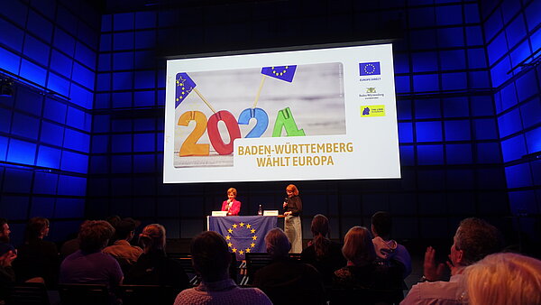 Foto einer Veranstaltung zum Europatag in einem modernen Konferenzraum. Zwei Sprecherinnen stehen vor einem Publikum, präsentiert auf einer Bühne mit einer großen Leinwand, die die Worte '2024 Baden-Württemberg wählt Europa' zusammen mit Europäischen Union Symbolen zeigt. 