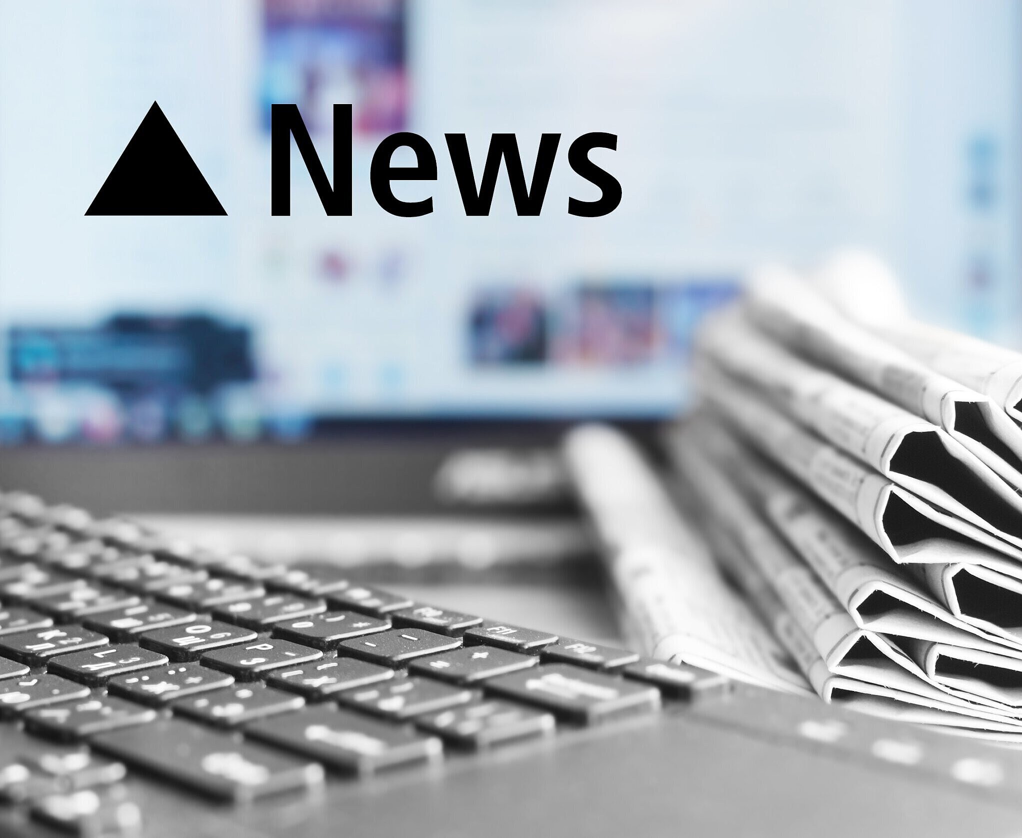 Ein Schreibtisch auf dem ein Stapel Zeitungen, eine Tastatur und ein Bildschirm zu sehen sind.