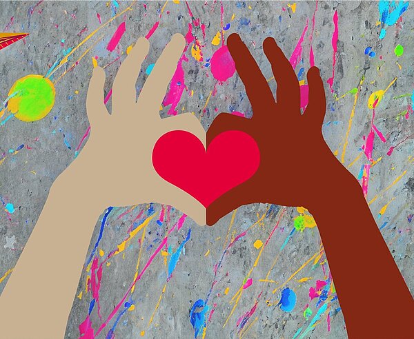 Eine dunkle und eine helle Hand formen mit Zeigefingern und Daumen ein rotes Herz. Hintergrund ist eine buntbekleckste Betonwand. Links oben ist der Kinderbüro Papierflieger zu sehen.